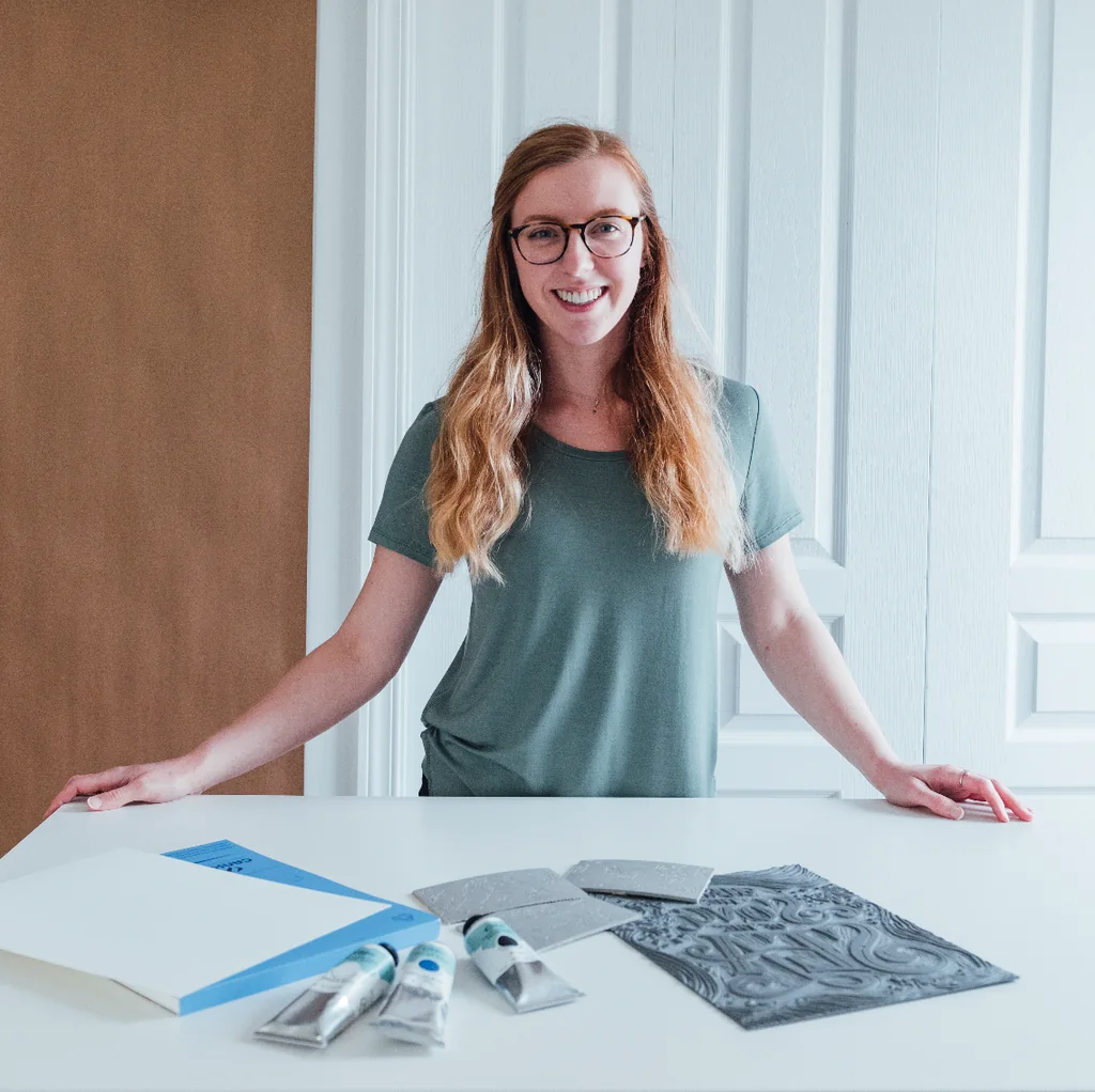 Kimber smiling at a table with her printmaking tools and artwork.