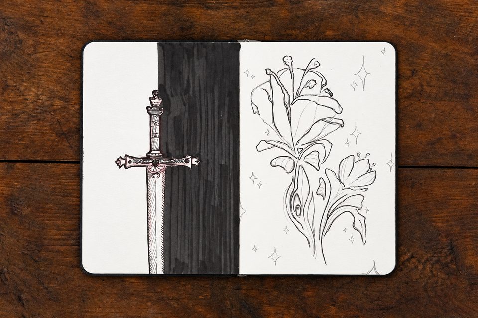 Open sketchbook on a wooden tabletop, with an ornate sword and flowing botanical ink drawings.